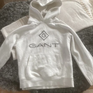 Vit hoodie från GANT - Säljer en stilren vit hoodie från GANT med deras ikoniska logga på bröstet. Tröjan har en klassisk känguruficka och en bekväm huva. Perfekt för en avslappnad stil.