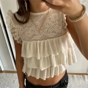 Beige spetsblus med volanger - Storlek S. Fin beige blus från Zara med spetsdetaljer och volanger. Den har en vacker broderad framsida och är ärmlös, vilket gör den perfekt för varmare dagar. Blusen har en romantisk och luftig design.