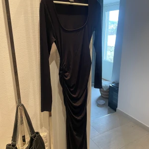 Svart långklänning från Gina Tricot - Svart långklänning från Gina Tricot med långärmad design och rynkdetaljer på sidan. Klänningen har en figurnära passform med urringning och är tillverkad i ett mjukt material. Den slutar lite nedanför vaderna på mig som är 172 cm. 