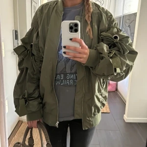 Olivgrön bomberjacka  - Snygg olivgrön bomberjacka med coola knytningar på ärmarna som gör jackan unik. Jackan har en dragkedja framtill och ribbade muddar vid ärmslut och nederkant. Perfekt till våren!
