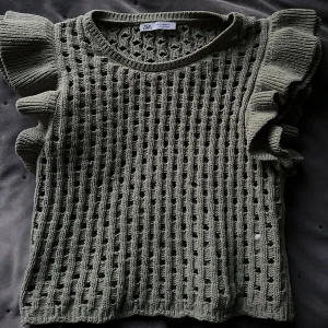 Olivgrön stickad topp från Zara - Säljer en olivgrön stickad topp från Zara med volangdetaljer på ärmarna. Toppen har ett hålmönster och är ärmlös, vilket gör den perfekt för varmare dagar.