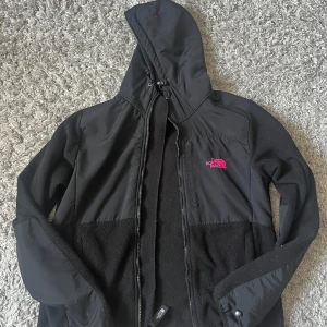 Svart fleecejacka från The North Face - Säljer en svart fleecejacka från The North Face med huva och dragkedja. Jackan har en broderad logga i rosa på bröstet. Perfekt för kyliga dagar.