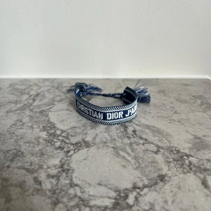 Blått armband från Christian Dior - Snyggt blått armband från Christian Dior med texten 'J'ADIOR'. Armbandet har en justerbar knytning och dekorativa tofsar i ändarna. Perfekt för att ge en stilren touch till din outfit.