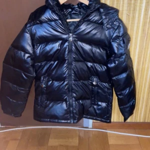 Svart pufferjacka från M Fashion - Snygg svart pufferjacka från M Fashion med dragkedja och knappar. Jackan har en huva och långa ärmar, perfekt för kyliga dagar. Den quiltade designen ger en modern look. Ett måste för garderoben!