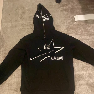 Svart hoodie med stjärnmotiv - Säljer en svart hoodie från Lukomachain med ett stort vitt stjärnmotiv och text på framsidan. Hoodien har dragkedja och en stor luca med texten 'Falling Star'. Perfekt för en cool och avslappnad stil. Inte använd mycket alls, skick 9/10