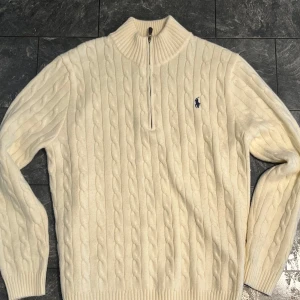 Krämit kabelstickad tröja från Ralph Lauren - Säljer en stilren krämvit stickad tröja från Ralph Lauren med kabelstickat mönster och en halv dragkedja vid halsen. Passar M och L