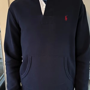 Mörkblå pikétröja från Ralph Lauren - Säljer en stilren mörkblå pikétröja från Ralph Lauren med klassisk krage och knappar. Tröjan har en broderad röd logotyp på bröstet och långa ärmar. Perfekt för en avslappnad men ändå elegant look. Är i jättebra skick och jätte snygg nu till sommaren!