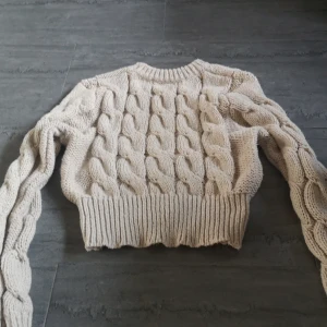Beige stickad tröja med flätmönster - Säljer en mysig beige stickad tröja med ett snyggt flätmönster. Tröjan är croppad och har långa ärmar med ribbade muddar. Perfekt för kyliga dagar!