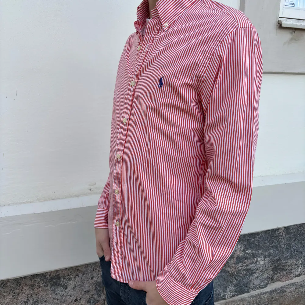Säljer en stilren röd- och vitrandig skjorta från Ralph Lauren. Skjortan har en slim fit passform och är långärmad med knappar framtill. Den har en broderad logotyp i blått på bröstet. Perfekt för en klassisk look.. Paidat.