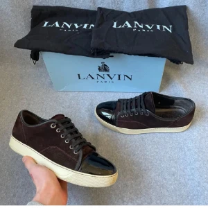 Vinröda Lanvins - Snygga vinröda sneakers från Lanvin med svart glansig tå och vita sulor. Skorna har snörning och är tillverkade i mocka med en stilren design. Perfekta för en trendig look. Box och en dustbag medkommer, tveka inte på att höra av er vid frågor!