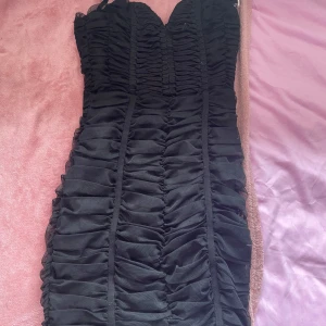 Svart klänning med rynkdetaljer - Snygg svart klänning med rynkdetaljer som ger en elegant look. Klänningen har smala axelband och en figurnära passform. Perfekt för en kväll ute eller en speciell tillställning.