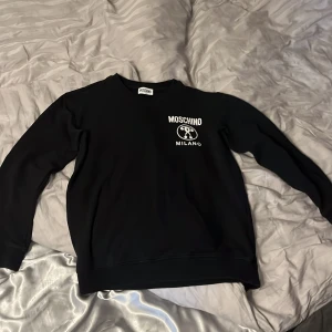 Svart sweatshirt från Moschino - Säljer en stilren svart sweatshirt från Moschino med vit logotyp på bröstet. Tröjan har en klassisk rund hals och långa ärmar. Perfekt för en avslappnad och trendig look. Nypris 2200 kr