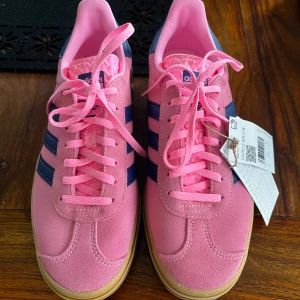 Rosa Adidas Gazelle Bold sneakers - Snygga rosa Adidas Gazelle sneakers med mörkblå ränder och klassisk gummisula. Skorna har snörning och är tillverkade i mocka för en stilren look. Perfekta för en färgglad touch till din outfit! Helt nya och köptes för 1400kr, säljs nu för 650 men kan diskuteras vid snabb affär 💞