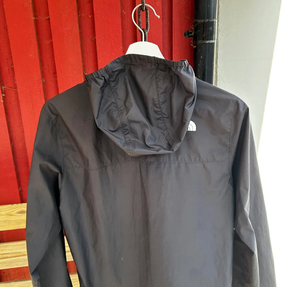 Säljer en svart vindjacka från The North Face med huva och dragkedja. Jackan har logotypen tryckt på bröstet och ärmen. Perfekt för blåsiga dagar. Ett litet hål vid vänster arm, men syns knappast.. Takit.