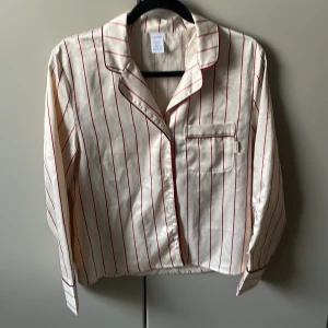 Vit och röd randig silkes pyjamas från Calvin Klein - Säljer en stilren vit pyjamas i silke med röda ränder från Calvin Klein. Setet består av en långärmad skjorta med knappar och en bröstficka samt matchande byxor. Perfekt för en bekväm natt med stil.