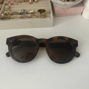 Bruna solglasögon i sköldpaddsmönster - Snygga solglasögon med rund form och klassiskt sköldpaddsmönster. De har en stilren design med mörka glas och små silverdetaljer vid bågarna. Perfekta för en solig dag!