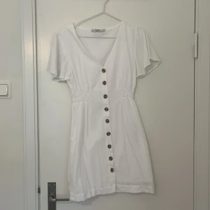 Vit kortklänning med knappar - Söt vit kortklänning med v-ringning och knappar framtill. Klänningen har kortärmade volanger och en smickrande passform med resår i midjan. Perfekt för varma dagar!