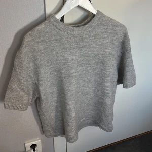 Grå stickad tröja från Gina Tricot i storleken M - Säljer en mjuk och bekväm grå stickad tröja från Gina Tricot. Tröjan har korta ärmar och en rund halsringning, perfekt för lager-på-lager under kyligare dagar.🩷