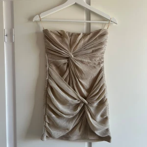 Beige axelbandslös klänning - Snygg guldig axelbandslös klänning med draperad design och knutdetalj framtill. Klänningen har en dragkedja bak och är perfekt för en elegant look.