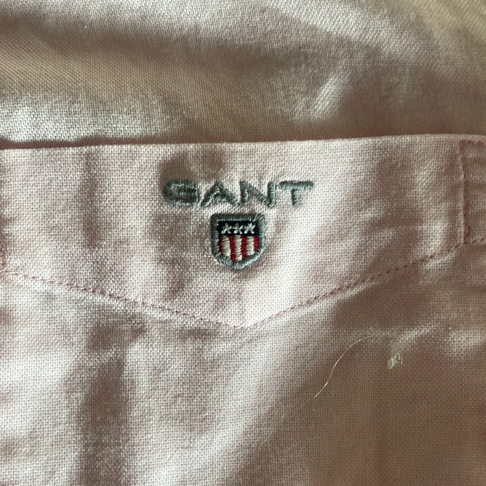 Säljer en stilren rosa skjorta från GANT med broderad logga på bröstfickan. Skjortan har långa ärmar och knappar framtill. Perfekt för en klassisk och avslappnad look.. Paidat.
