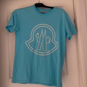 Turkos t-shirt från Moncler - Säljer en snygg turkos t-shirt från Moncler med ett stort vitt logotryck på framsidan. T-shirten är kortärmad och gjord i mjukt material, perfekt för en avslappnad stil. Passar bra till både jeans och shorts!