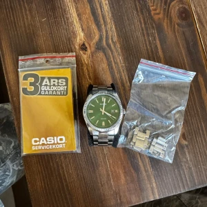 Grön klocka från Casio - Säljer en stilren Casio-klocka med grön urtavla och silverfärgad boett. Klockan har ett datumfönster och kommer med extra länkar samt ett servicekort med 3 års garanti. Perfekt för dig som gillar en klassisk look med en twist.