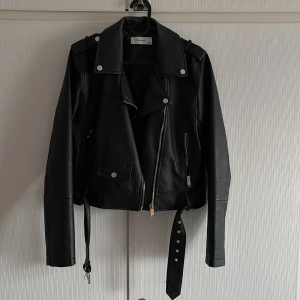 Svart skinnjacka med dragkedja  - Snygg svart skinnjacka från Zara med dragkedja och knappar. Jackan har långa ärmar och ett bälte i midjan för en cool look. 