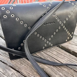 Svart axelremsväska från Ulrika! - Svart axelremsväska med snygga detaljer och många fack från svenskdesignade Ulrika. Mått: 20 x 25 cm. I skick som ny!