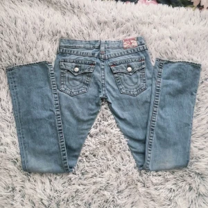 True Religion Low-waist Bootcut Jeans - Eftertraktade True Religion jeans med detaljerade fickor med lock. Jeansen har även röda små broderingar/detaljer. OBS! Finns någon liten fläck på sista bilden men den är så liten att man knappast märker av den. Innerbenslängd: 83cm och midjemått: 38cm rakt över. Storlek 28 men skulle säga att de är mindre i storleken och inte jätte stretchiga så passar 26-27 bäst.