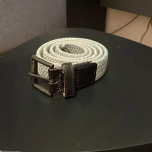 Beige flätat bälte med läder spänne - Snyggt beige flätat bälte med ett robust mörkbrunt spänne och detaljer i skinn. Perfekt för att ge en stilren touch till din outfit.