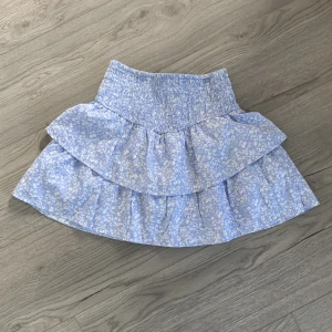 Blommig blå kjol med volang - Söt blå kortkjol med blommigt mönster och volangdetaljer. Perfekt för sommarens varma dagar. Den har en elastisk midja för en bekväm passform.