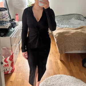 Svart långärmad klänning med slits - Elegant svart klänning med lång ärm och djup v-ringning. Klänningen har en smickrande slits framtill och en knutdetalj i midjan som ger en fin silhuett. Perfekt för en kväll ute eller en dag på stranden.
