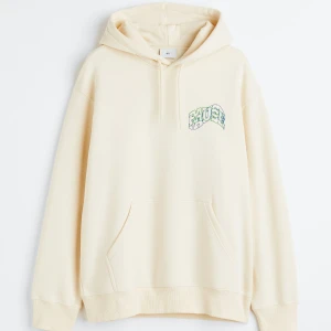 Beige hoodie från H&M - Säljer en beige hoodie från H&M med textdetalj i grönt och blått. Den har en avslappnad passform och en klassisk känguruficka framtill. Perfekt för en casual look.