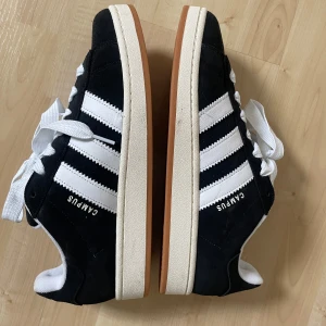Svarta Adidas Campus sneakers - Snygga svarta Adidas Campus sneakers med vita ränder och snörning. Skorna har en klassisk design med en bekväm passform och en gummisula i kontrasterande färg. Aldrig använda, bara testade i affär!  Köptes i USA, så jag har tyvärr inte kvittot eller boxen kvar! Storlek: 44 2/3 Supersköna, snygga, trendiga, passar till allt, bra i storleken, unisex! Hör av er vid frågor💕 