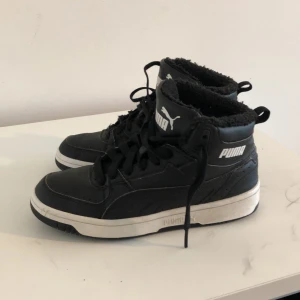 Svarta sneakers från Puma - Snygga svarta sneakers från Puma med hög krage och snörning. Skorna har en vit sula och Puma-loggan synlig på sidan och tungan. Perfekta för en sportig look.