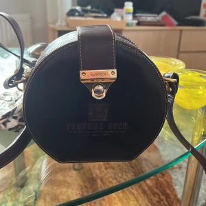 Rund brun axelväska från Tortue Duck - Säljer en unik rund axelväska i mörkbrunt skinn från Tortue Duck. Väskan har en justerbar axelrem och ett snyggt metallspänne framtill. Perfekt för att ge din outfit en cool touch.