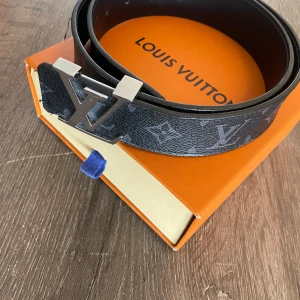 Svart bälte från Louis Vuitton - Snyggt svart bälte från Louis Vuitton med det ikoniska LV-mönstret och en stilren silverfärgad spänne. Perfekt för att ge din outfit en lyxig touch. 