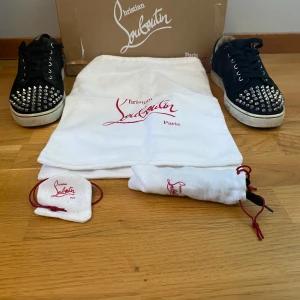 Louboutin skor - Snygga svarta sneakers från Christian Louboutin med ikoniska röda sulor. Skorna har en rund tå och är dekorerade med nitar på tån och hälen. De har snörning framtill. Allt kommer till och har kvitto