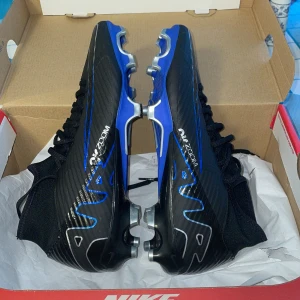 Nike Zoom Mercurial fotbollsskor - Snygga Nike Zoom Mercurial fotbollsskor i svart och blått med vit logga. Skorna har snörning och en strömlinjeformad design för optimal prestanda på planen. Perfekta för fotbollsspelare som vill ha både stil och funktion. 