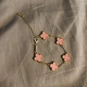 Guldarmband med fyrklövermotiv - Charmigt guldarmband med fyrklövermotiv i rosa och vitt. Armbandet har en justerbar kedja och är perfekt för att lägga till en touch av elegans till din stil. Pris är för ett