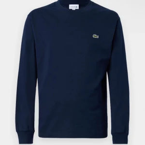 Mörkblå långärmad tröja från Lacoste - Säljer en stilren mörkblå långärmad tröja från Lacoste med det klassiska krokodilemblem på bröstet. Perfekt för en avslappnad och snygg look. Tröjan har en rund halsringning och ribbade muddar vid ärmslut.