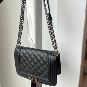 Svart quiltad väska med kedja - Snygg svart axelväska från Ginatricot med quiltat mönster och kedjedetaljer. Väskan har en praktisk dragkedja inuti och en elegant låsdetalj framtill. Kedjan går att justera och ha crossbody.