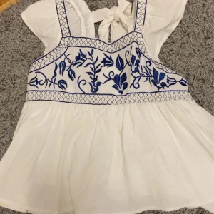 Vit blus med blå broderier - Charmig vit blus med vackra blå broderier och knytband i ryggen. Blusen har breda axelband och en luftig passform, perfekt för varma dagar.