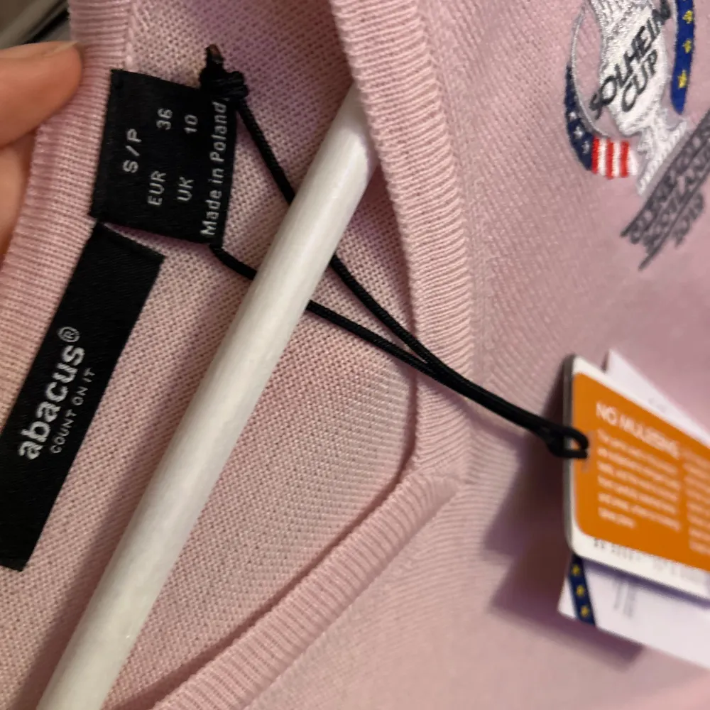 Säljer en ljusrosa tröja från Abacus med Solheim Cup 2019 broderi på bröstet. Tröjan har en v-ringad design och långa ärmar, perfekt för golfentusiaster. Tillverkad i ett mjukt material för komfort.. Neuletakit & Villapaidat.