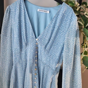 Vacker Adoore klänning i blått. Storlek M - Blå klänning från märket Adoore. Storlek M. I superfint skick. Knappar fram på klänningen. Resår vid ärmsluten. Fin rynk-detalj på ryggen. Denna klänningen är mindre i midjepartiet och Adoore rekommenderar att du går upp 1 storlek. Så om du normalt är en storlek S så borde denna passar dig. Alla är vi olika men det är vad Adoore rekommenderar.