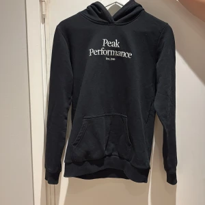 Svart hoodie från Peak Performance - Stilren svart hoodie från Peak Performance med vit logga på bröstet. Tröjan har en klassisk känguruficka och en bekväm huva. Perfekt för en avslappnad look. ordinarie pris (799kr). Juniorstorlek 170. Lite använd och mycket bra skick! 