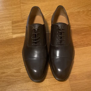 Capo oxfordkostymsko i både svart och mörkbrunt kalvskinn – handgjord i Italien - Säljer en oanvänd klassisk oxfordkostymsko från Capo i mörkbrunt kalvskinn med dekorativ tåkappa. Handgjord i Italien med nappafoder, vadderad innersula och durksydd lädersula i “Channel”-konstruktion. Elegant, bekväm och perfekt till mörk kostym. Storlek 39 men passar 40 Nypris: 4500kr Mitt pris: 1500kr