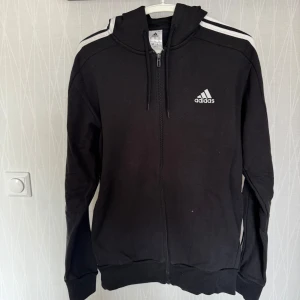 Svart hoodie från Adidas - Säljer en svart hoodie från Adidas med dragkedja och vita ränder längs ärmarna. Den har en justerbar huva och ribbade muddar. Perfekt för en sportig look eller avslappnad stil.