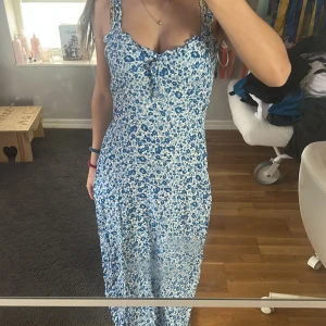 Blommig långklänning från FBSister - Säljer en vacker blommig långklänning från FBSister i blått och vitt. Klänningen har en öppen rygg och en slits framtill. Den är ärmlös med knytning vid bysten, perfekt för sommarens varma dagar.