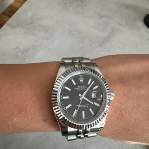 Silverfärgad klocka R*lex - Säljer en elegant silverfärgad klocka med svart urtavla och datumvisning. Klockan har en räfflad bezel och en länkarmband i rostfritt stål. Perfekt för den som söker en tidlös accessoar.